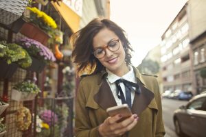 A smiling woman uses her phone outside a colorful urban flower shop on a sunny day.